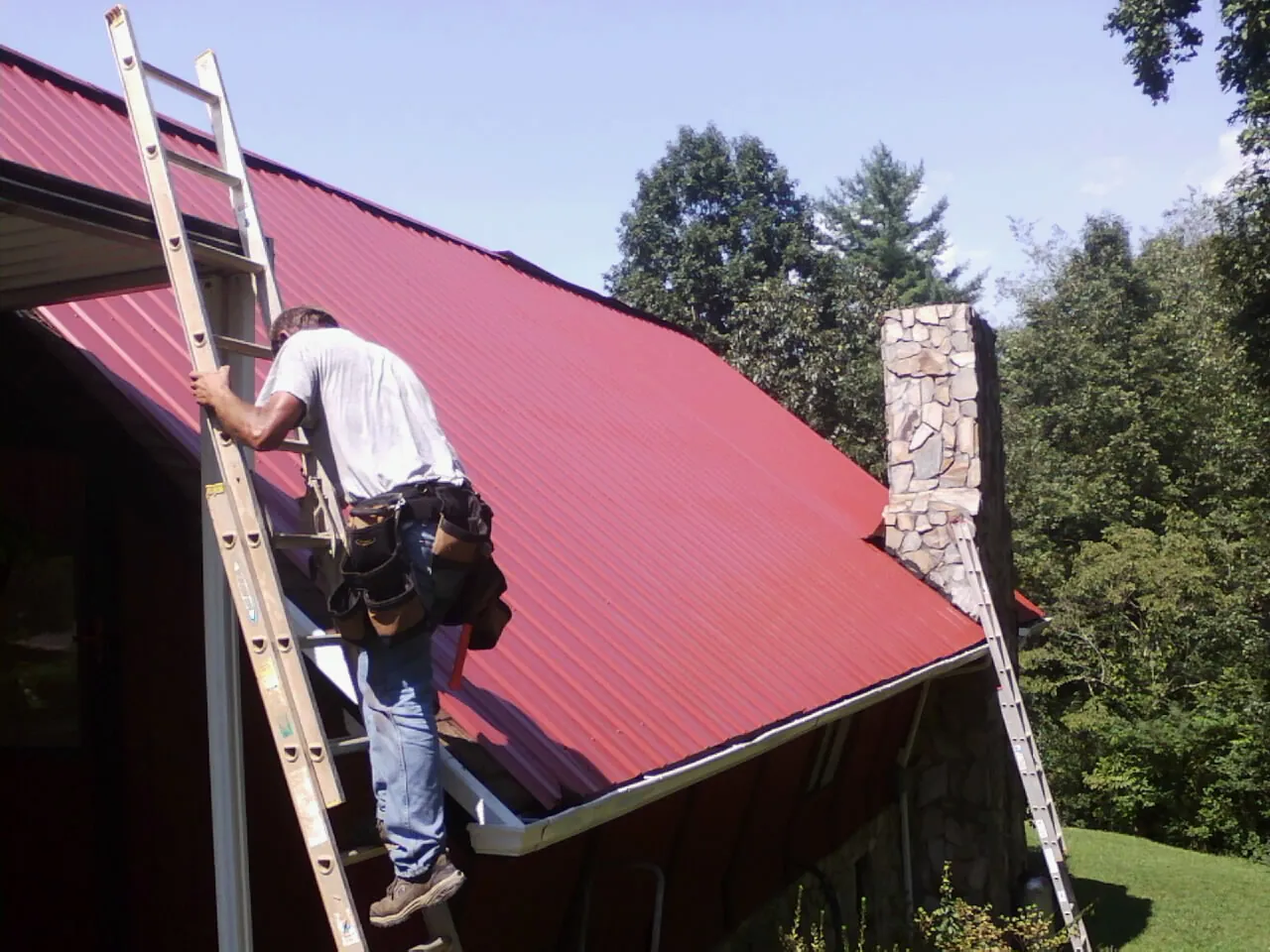 Roofer on a ladder beside a red metal roof and stone chimney — Soffit & Fascia Repair by Roofing Brothers in Trumbauersville